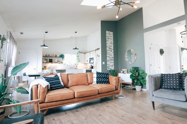 Open-plan living room with tan leather sofa, teal accent wall, pendant lights, and kitchen visible in background