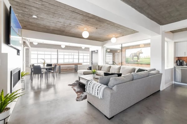 Open-plan living room with gray sectional sofa, concrete floors, wood ceiling beams, and large windows overlooking pasture.